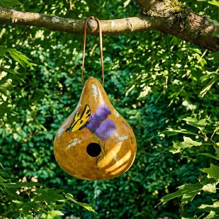 A hand-painted butterfly gourd birdhouse hanging securely from a high tree branch, surrounded by lush green foliage and a garden in the distance, positioned safely away from ground predators.